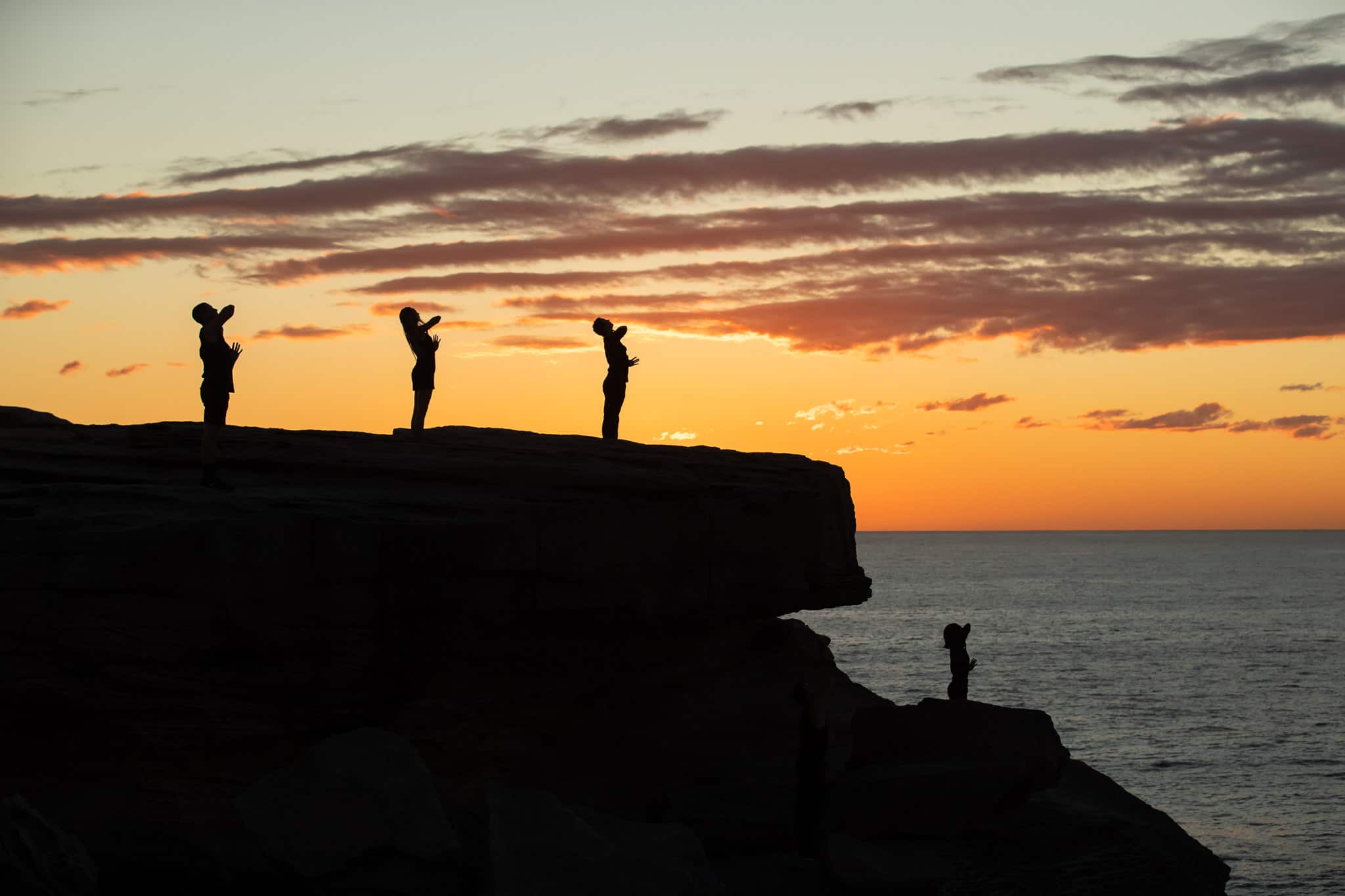 Four dancers stand with their arms outstretched overlooking the ocean on a rocky cliff at sunrise.