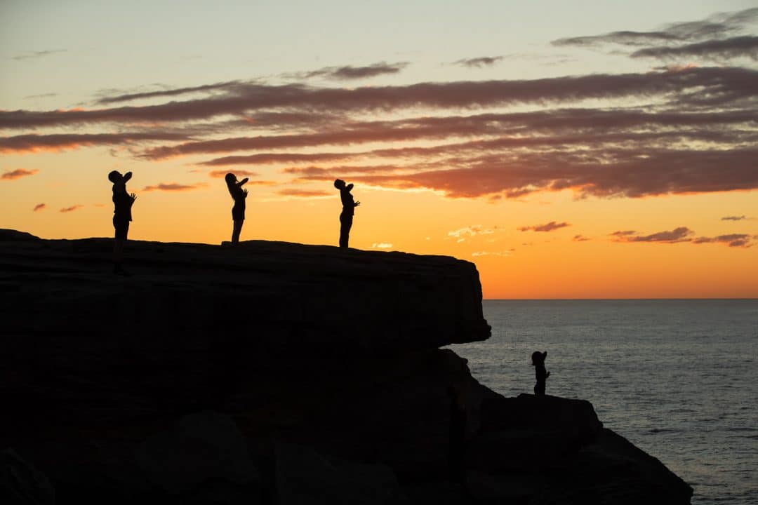 Four dancers stand with their arms outstretched overlooking the ocean on a rocky cliff at sunrise.