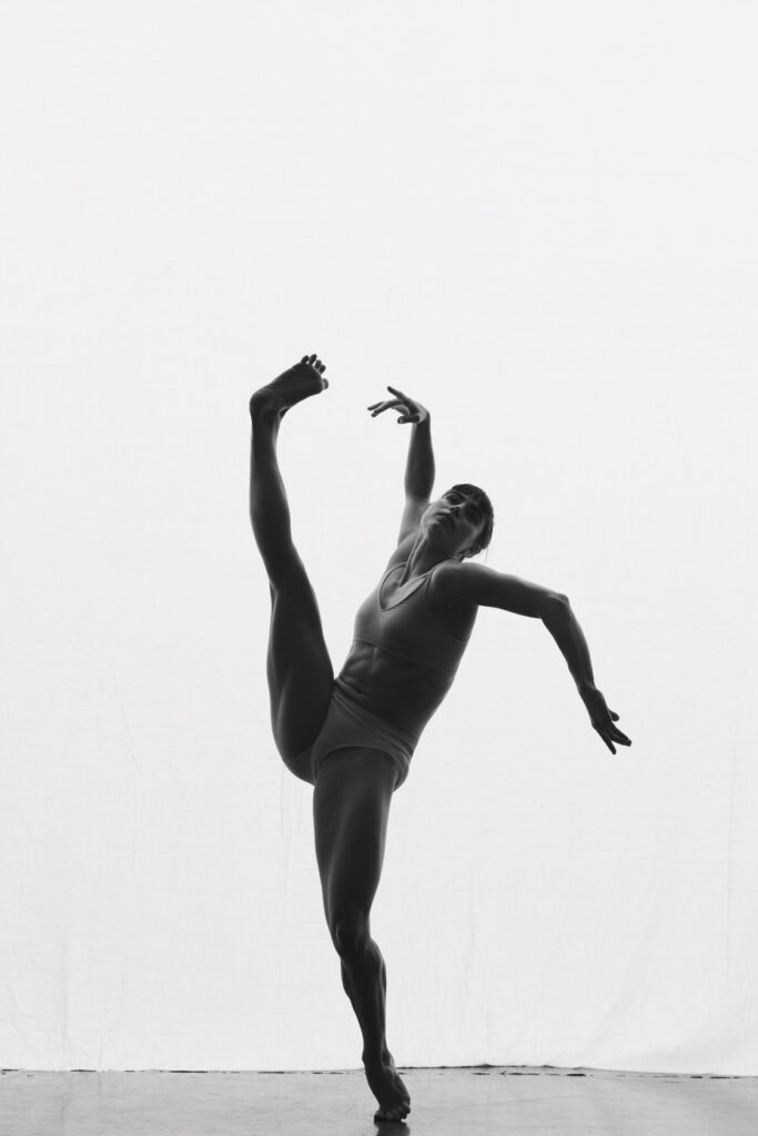 A female dancer kicks a leg to the sky and holds her arms outstretched.