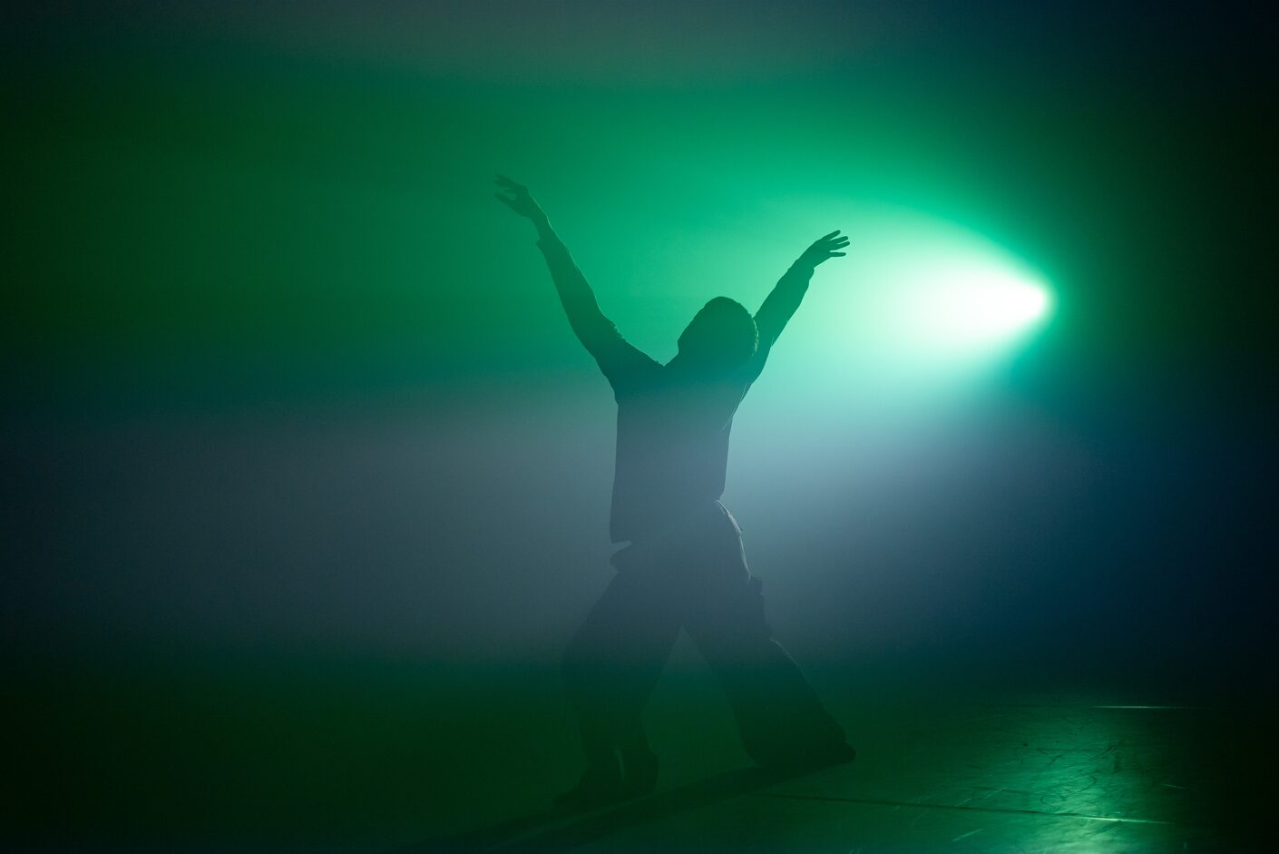 Dancer with their back to us and arms raised, bathed in a greenish light, sourced to their right.
