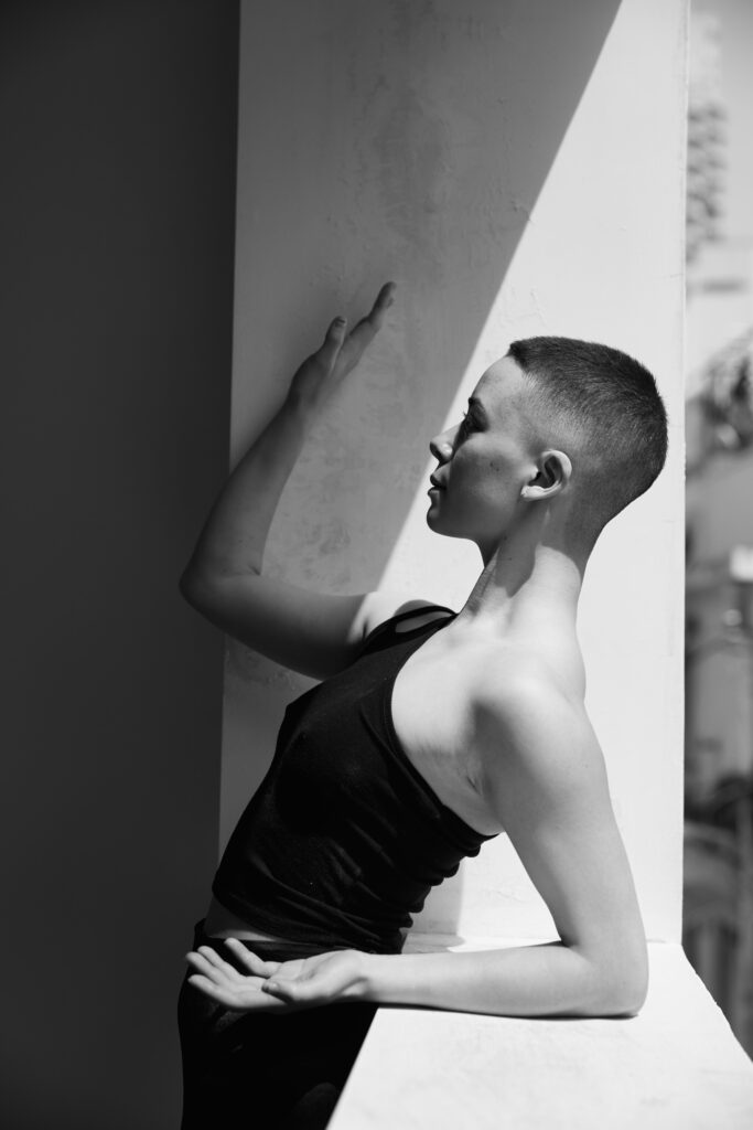 A female dancer stands side on, leaning backwards with her arm bents and palms facing the sky.